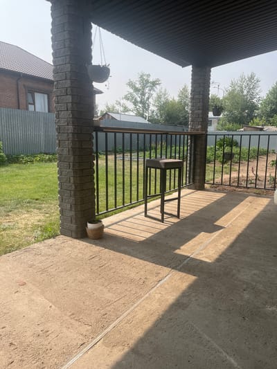 This is a horizontal, eye-level shot of a covered porch with brick pillars. In the foreground, a concrete floor is visible, casting long shadows from the porch roof. A dark, minimalist stool sits on the porch, facing away from the camera. Behind the stool, a black metal railing encloses the porch, beyond which is a grassy area and a metal fence.
Two brick pillars support the roof, one in the left foreground and another further back. Hanging from chains attached to the porch roof are two dark planters, one with a visible plant. To the right of the further pillar, a woven basket containing a white pot sits on the concrete.
In the background, glimpses of buildings with dark roofs and grey metal siding can be seen, along with lush green trees. The sky is clear and bright, suggesting a sunny day. The scene appears to be a residential outdoor area, possibly a backyard or patio. The image lacks any people or animals, and there is no visible text. The time of day appears to be daytime, likely late morning or early afternoon due to the strong sunlight and shadows.