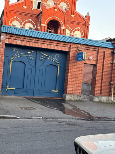 This media file captures a scene in Sulak, Russia, featuring the exterior of a brick building with a prominent blue gate. The building has an ornate red brick facade with arched windows, suggesting a historical or decorative architectural style. Above the gate, a blue sign with white lettering reads "Смоленский HOSTEL," indicating it is a hostel. The gate itself is a dark blue, featuring decorative golden scrollwork and vertical lines.
To the right of the building and gate, a paved road is visible. A small section of a white vehicle is present on the far right edge of the frame. The road shows signs of wear, with cracks and a dark, wet stain that appears to be a spill or leak. A manhole cover is also visible on the road. The sky is overcast, suggesting a cloudy day. The lighting is diffused, indicating it is likely daytime, but the overcast conditions prevent strong shadows. The overall impression is of an urban or semi-urban setting, possibly residential or commercial, given the presence of the hostel sign. There are no people visible in the frame.
