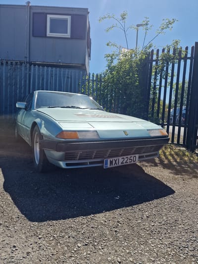 A light blue or teal classic car, possibly a Ferrari based on its angular design and small badge on the bonnet (though the badge itself is indistinct), is prominently featured in this outdoor image. The car is parked, showcasing its front and front-right side, with its polished surface reflecting the bright blue sky above. A UK license plate, clearly visible on the front, reads "WXI 2250" with the "GB" identifier.
The setting appears to be an industrial or commercial area in South Tyneside, United Kingdom, under bright, clear conditions. To the left of the car, a two-story structure resembling stacked portable cabins or shipping containers is visible, predominantly gray-blue, with a white-framed window on the upper unit. Behind the car and extending across the background is a dark metal fence, likely security fencing, entwined with green foliage and branches. The ground is a mix of gravel and asphalt, with a distinct, long shadow cast by the car, indicating strong sunlight, likely in the mid-morning or late afternoon. No people or specific activities are visible; the scene captures a static moment on a sunny day.
