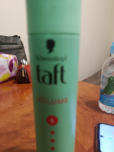 The image displays a close-up view of a green aerosol can for hairspray, specifically "Taft Volume" by Schwarzkopf. The can is positioned prominently in the foreground, slightly out of focus. To the left of the can, partially visible, are two cosmetic bags or pouches, one with a floral pattern in pink, purple, and yellow, and another smaller, reddish-brown textured bag with a gold zipper pull. In the background, a dark gray bag with multiple zippered compartments is visible.

To the right of the hairspray can, a plastic bottle of "Sagua Woda Zródlana Niegazowana" (Sagua Natural Still Water) is present. The bottle has a blue label with an illustration of a green bird or gecko. Below the water bottle, the corner of a smartphone is visible, displaying a bright screen with some icons.

The setting appears to be indoors, on a polished wooden surface, likely a table. The lighting is artificial and somewhat diffused. There are no explicit cues about the time of day or weather. The overall impression is one of a casual arrangement of personal items. The visible text includes "Schwarzkopf," "taft," and "VOLUME" on the hairspray can, and "Sagua Woda Zródlana Niegazowana" on the water bottle. The location context provided is Khmilnyk, Ukraine.