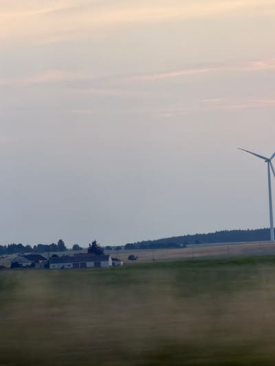 This is a distant shot of a rural landscape, likely at dusk or dawn. In the mid-ground, a cluster of white farm buildings with dark roofs is visible. To the right of the farm buildings, a large white wind turbine stands tall against the pale sky. The foreground is a blurred field of grass, suggesting motion, possibly captured from a moving vehicle. A dark line of trees forms the horizon behind the farm. The sky is a pale pink and grey, with wispy clouds. The image does not contain people, actions, or interactions. The location is identified as being in the vicinity of Burkat, Poland.