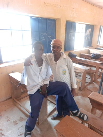 The scene is a classroom in Mada, Nigeria. Two young males are centrally positioned. The male on the left, wearing a white long-sleeved shirt, white undershirt, and dark blue trousers with black and white sandals, is seated on a wooden desk. He looks forward with a slight smile. The male on the right, wearing a light cream long-sleeved shirt with a school emblem on the left chest, dark blue trousers, a light brown cap, and brown sandals, stands behind the desk, his right arm resting on the left male's shoulder. He looks directly at the camera. The classroom contains multiple rows of wooden student desks and benches, some marked with "ACHIEVE C" and "C1020". The light-colored walls display various chalk or paint graffiti, including "HEAVEN," "ZINO vs D. DOLLCE," and "ROBOBO vs EMPTIE." Large windows with dark blue frames and security grates are present on the left wall, admitting bright natural light. Two dark bags are visible on desks in the background. The floor is a worn, light-colored concrete.