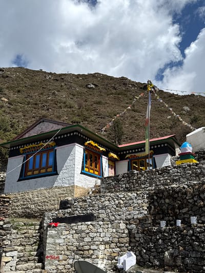 The image captures a vibrant traditional building in Khumjung, Nepal, set against a rugged, rocky mountainside under a partially cloudy, blue sky. The multi-story building features white stucco walls adorned with colorful trim in black, blue, yellow, red, and green. Its windows have brightly painted wooden frames in shades of yellow, blue, and red.
A tall flagpole stands prominently in front of the building, decorated with numerous multi-colored Buddhist prayer flags (lungta) that extend towards the mountain in the background. Extensive dry-stone walls form the building's foundation and surrounding structures, reflecting traditional Himalayan construction.
On the right side of the image, a tall stone wall features several small, white rectangular objects and a piece of red fabric. A small, grey satellite dish is mounted near this wall. The scene is illuminated by bright daylight, casting clear shadows.
No people are visible, but the presence of prayer flags and the well-maintained architecture indicate a local settlement, likely a guesthouse or residence. On a stone block on the right, the word "TOILET" is visible in red paint, accompanied by an arrow pointing right, indicating the location of a facility. The setting is characteristic of a high-altitude Himalayan village.