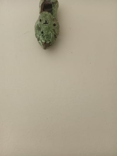 The image shows a green ceramic or stone object, possibly a decorative piece, affixed to an off-white wall. The object has a textured surface with several holes and an opening at one end. The wall appears to be part of an interior space. The specific location in Ksar-el-Kebir, Morocco, cannot be determined from the image alone. There are no people visible in the image.
