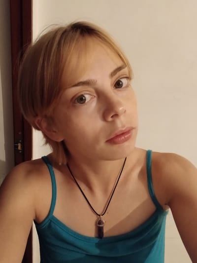 The image is a close-up, waist-up, portrait of a young Caucasian female with blonde hair and light skin. She is wearing a turquoise tank top and a dark necklace with a purple pendant. The background consists of a plain, light-colored wall and a dark wooden door frame on the left side of the image. The lighting suggests an indoor setting. There is no indication of the specific location within the city of Taraz, Kazakhstan.