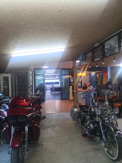 The image captures the interior of a motorcycle-themed establishment in Surat Thani City Municipality, Thailand. Two motorcycles, one red and one black, dominate the foreground. Behind the black motorcycle, a counter with two seated people (a woman and a man), can be seen. A third person stands near the counter. A series of framed motorcycle-related posters line the upper wall, with hanging lights. A doorway or opening leads to an outside area where a white van is parked. The interior ceiling appears to be constructed from plywood.