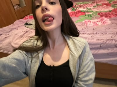 The scene depicts a female subject indoors, identified as being in Волховец, Russia. The subject, viewed from a low angle, occupies the majority of the frame, presenting with long, dark hair. She is clad in a grey zip-up hooded sweatshirt worn over a black button-front top. Her mouth is open, with her tongue extended forward. In the background, a bed is visible, covered by two distinct blankets: the upper portion features a floral pattern of pink and red blossoms, while the lower portion is a light purple with white script detailing. A light brown wooden wardrobe or cabinet is partially observable to the left of the bed. The foreground consists of a solid, light-colored floor surface.