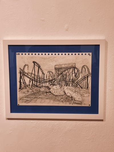 The image shows a framed black and white drawing of an abandoned amusement park with a roller coaster as a central element. The drawing is on paper with perforation at the top, suggesting it was torn from a notebook. The roller coaster structure, with the sign reading "ROLLER COASTER", is surrounded by the wreckage of cars and other park elements, giving the impression of ruin and decay. The drawing is mounted within a blue border and then placed inside a white frame, which is hanging on a white wall. The scene is not recognizable as a specific location in Querétaro, Mexico, as it appears to be a generic abandoned amusement park. There are no people visible in the scene.