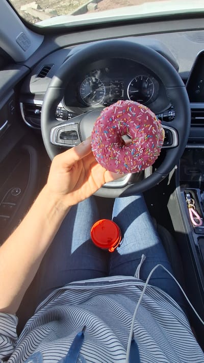 The image captures a first-person perspective from inside a car, likely in Яльгелево, Russia, on a sunny day. A person, wearing blue jeans and a blue and white striped shirt, is holding a bright pink donut covered in colorful sprinkles. Their right arm and hand are prominently displayed, illuminated by direct sunlight, creating distinct shadows.
The car's interior features a black steering wheel, behind which a digital and analog dashboard is visible. The digital display shows "10:32" (time) and "18.3km" (distance). The analog speedometer indicates 0 km/h, and the tachometer is also at 0, suggesting the car is stationary. Other dashboard elements include fuel and temperature gauges. Buttons on the steering wheel include "SET," "RES," "CRUISE," and "MODE." To the right of the steering wheel, part of the center console can be seen, with a pink keychain and other small items. A red lidded container, possibly a small cup or bottle cap, rests on the person's lap. A white cable is draped across their leg and the striped shirt. The overall scene suggests a break or a snack taken while parked.