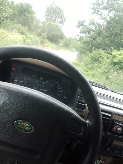 The image offers a driver's perspective from inside a Land Rover, looking out through a rain-splattered windshield onto a lush green, overgrown landscape, indicative of a rural area like Zemen, Bulgaria. The steering wheel, prominently featuring the "LAND ROVER" emblem and "SRS AIRBAG" text, occupies the lower left.
A portion of the car's dashboard is visible behind the steering wheel, displaying a speedometer at 0 km/h and a tachometer showing an engine speed below 1000 r/min, suggesting the vehicle is stationary or idling. Several warning lights are illuminated, including the amber "ABS" (Anti-lock Braking System) light, a red brake warning light (a circle with an exclamation mark), and an amber "TC" (Traction Control) light. Other interior elements include various control buttons on the steering wheel and part of the center console with radio controls, including an "AUTO" button.
Outside the vehicle, a dense expanse of green trees, bushes, and tall grass dominates the view. A narrow, unpaved or dirt road is faintly visible winding through the vegetation in the distance. The overcast sky and water droplets on the windshield indicate either current rainfall or recent wet weather, contributing to the natural, verdant ambiance. The scene depicts a daytime setting, possibly during or after a rain shower, suggesting an off-road excursion or travel through a natural environment.
Visible text includes: "LAND ROVER", "SRS AIRBAG", "r/min x1000", "km/h", "ABS", "TC", and "AUTO".