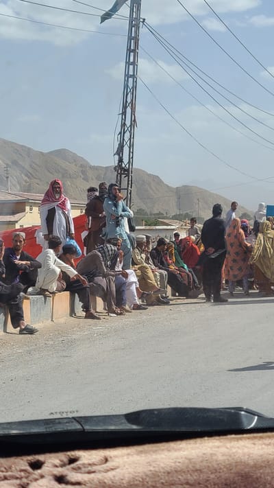 The image depicts an outdoor scene along a paved road in a mountainous region, likely Kuchlak, Pakistan. In the background, rugged, light-colored mountains rise under a bright, clear sky with scattered clouds. A prominent utility pole with numerous power lines stands on the left side of the road.

A large group of people, mostly men, are gathered along the roadside. Many are seen sitting or lying down on the ground, on curbs, or on elevated steps, suggesting a period of rest or waiting. Their attire is traditional, consisting primarily of shalwar kameez, with many men wearing turbans or headscarves. A few women, also in traditional dress, are visible further back among the crowd. Some individuals are standing, observing the scene. The photograph appears to be taken from inside a vehicle, with a portion of a car's interior visible on the far right.

The strong, direct sunlight casting sharp shadows, particularly from the utility pole, indicates it is midday or early afternoon. The environment appears dry and dusty. The people generally seem calm and at ease, perhaps tired or resigned, rather than actively engaged in an urgent activity.

On the paved road, faint white markings are discernible, including text that appears to read "ATDYOT," followed by another faint inscription that looks like "TOYOTA" on a dark, possibly recessed, object.