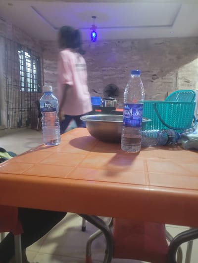 The scene appears to be indoors in Ogbunike, Nigeria, featuring an orange plastic table with several bottles of water (brands: 'KP Table Water' and 'Afinia Aqua') placed on it. A metal pot rests on the table next to one of the water bottles. A person wearing a light pink shirt and dark pants can be seen in the background. Other items include a blue plastic basket, and a fan mounted on the wall. The room appears to be lit by fluorescent lighting, including a purple decorative light fixture. The background shows a wall with a textured or worn appearance and a barred window. Part of a red and silver chair can also be seen on the right side of the table.