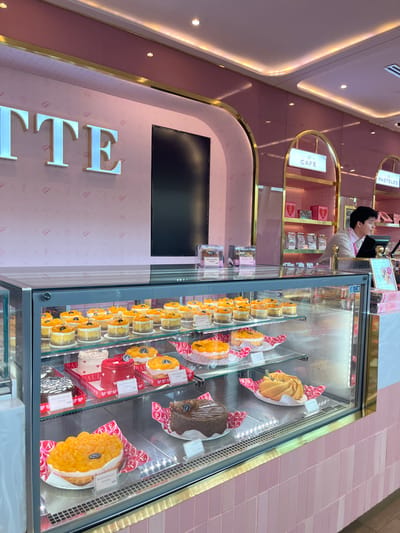 A brightly lit café with a display case filled with small cakes, tarts, and pastries. A person stands behind the counter. The café features pink walls, gold accents, and a TV screen. The name of the café is vertically spelled out in large, silver letters. Pastries are individually displayed in the refrigerated case. The general aesthetic is modern and clean.