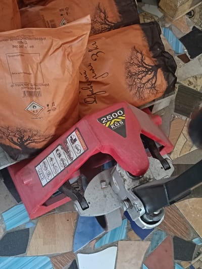 The image shows a red hydraulic pallet jack positioned on a floor made of colorful, irregular ceramic tiles. The pallet jack has a label with "2500 KGS CAPACITY" printed on it. Behind the pallet jack are two large, brown bags. One bag, on the left, has text printed on it, including "СОСТАВ: ДРЕВЕСИНА ЛИСТВЕННЫХ ПОРОД" and "ВЕС ЗКТ +/- 10%". It also features a barcode and several hazard symbols, including a flame symbol with "4.2" and a symbol for flammable solids. The other bag, on the right, has a design of a silhouetted tree against an orange background. The scene is illuminated by artificial light, casting shadows. There are no people visible in the image. The setting appears to be an indoor storage area or workshop. No specific location in Berdiansk, Ukraine, can be identified from the visual information alone.
