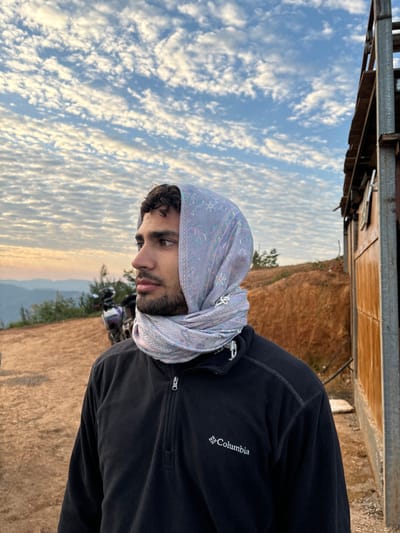 A person stands outside with a scarf on their head. They wear a black Columbia fleece jacket. The background shows a landscape with a cloudy sky, a hill, and some greenery. Part of a building is visible in the upper left corner. The photo is taken at a slight angle.