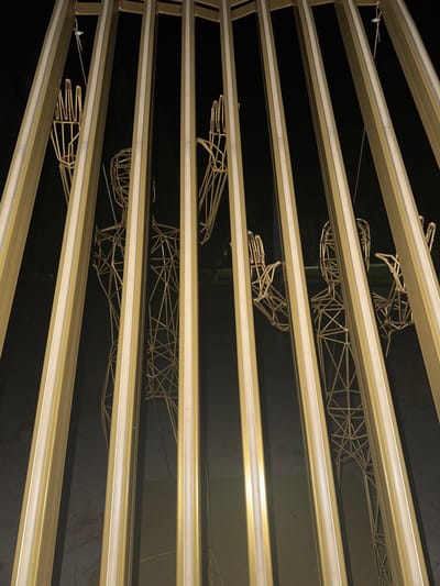 The image shows a close-up, angled view of several golden wireframe sculptures of human figures. These figures are suspended and viewed from above, appearing to be part of a larger installation. They are positioned behind horizontal golden bars with embedded white lights. The background is dark, suggesting the photo was taken at night or in a dimly lit indoor space. The wireframe construction of the figures is intricate, showing skeletal outlines of torsos, arms, and legs. Some figures appear to be reaching out or in motion. The lighting creates a reflective effect on the golden bars and highlights the geometric shapes of the sculptures. There are no visible text elements in the image. The setting appears to be an outdoor display or an exhibition space.