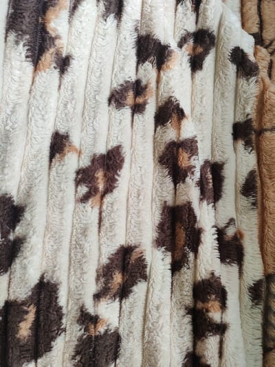 The image is a close-up, overhead view of a textured, plush fabric with a leopard print pattern. The fabric is predominantly off-white with irregular splotches of dark brown and lighter tan. It has a ribbed texture with raised vertical lines running through it. The lighting is even, and there are no shadows to indicate the direction of light. The scene appears to be a flat surface, possibly a blanket or upholstery, indoors. There is no indication of the city of Первомайский, Russia in this image.