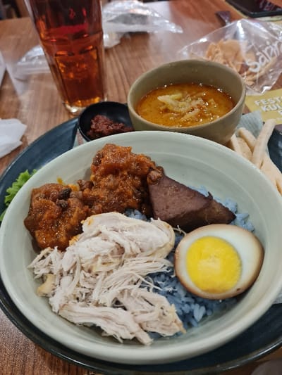 The scene depicts a prepared meal set on a light-colored wooden table within a dining establishment. The central object is a white bowl with a dark rim, resting on a dark plate, containing blue rice. Arranged on the rice are shredded white chicken, a portion of reddish-brown stewed meat, a dark brown, triangular piece of protein, and a halved hard-boiled egg with a dark-edged white and prominent yellow yolk. A green lettuce leaf is visible beneath the bowl.
Accompanying the main dish are two smaller bowls: a black bowl holds dark red chili paste, and a light brown bowl contains an orange-yellow curry or soup. A tall glass containing an amber liquid, consistent with iced tea, stands to the left.
In the background, a clear plastic bag containing flat crackers is visible, bearing the name 'Sarina'. A partially visible yellow sign displaying the text 'FESTIV KULI' is also present. The 'Sarina' branding and 'Festival Kuliner' sign indicate the location is within or proximate to the Sarinah department store and cultural center, situated on Jalan M.H. Thamrin, Menteng, Central Jakarta, Special Capital Region of Jakarta, Indonesia.