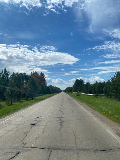 The image displays a long, straight asphalt road extending into the distance under a bright blue sky. The road surface is heavily cracked and shows considerable wear, with a faded white line marking its right edge.
To the left of the road, a dense line of tall green trees forms a border, suggesting a forest or woodland. Partially visible through and above these trees, running parallel to the road, is a prominent silver-colored pipeline, reflecting the sunlight. To the right of the road, there is a strip of green grass, speckled with small yellow flowers.
The sky above is clear and a vibrant blue, filled with numerous scattered white clouds. Some clouds are wispy and streaky, while others are more defined and puffy, particularly towards the horizon. The bright lighting indicates it is daytime, likely mid-morning or afternoon, with strong sunlight casting minimal shadows.
No people, vehicles, or specific activities are depicted in the scene. The setting appears rural or remote, consistent with the provided location context of Belorechensky, Russia. There is no visible text in the image.