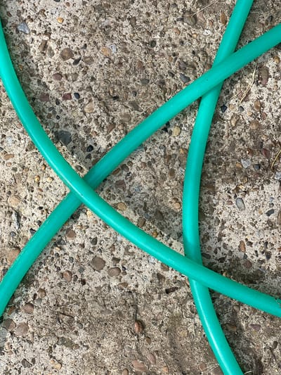 A bright green garden hose lies coiled and crossed over a concrete surface speckled with small, rounded pebbles of various earth tones. The concrete appears weathered and slightly uneven. The lighting suggests it is daytime, with shadows subtly highlighting the texture of the hose and the ground. There are no people or animals visible in the image. The scene is a close-up, overhead view of the hose and pavement, emphasizing the contrast in textures and colors. The location within Yakshur-Bod'ya, Russia is noted as an outdoor setting, likely a yard or driveway.
