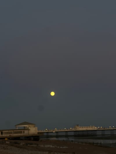 This image depicts Worthing Pier in Worthing, United Kingdom, at dusk or night. A prominent, bright yellow moon is visible in the upper-middle portion of the dark grey sky. The pier extends from the left side of the frame into the background, showcasing its architectural structures. A multi-story, light-colored pavilion with a pitched roof is visible at the shore end of the pier on the left. The main length of the pier is illuminated by a series of small, artificial yellow lights along its railing, which cast faint reflections on the wet foreshore below. The foreground consists of a dark, rocky and sandy beach, indicating a low tide, with several indistinct human figures walking on the exposed wet sand beneath the pier structure. The overall environment is calm, with a clear sky allowing visibility of the moon and artificial lights providing illumination to the pier and its immediate surroundings.