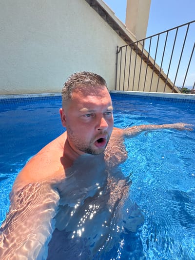 A man is pictured waist-deep in a swimming pool with clear blue water. He is facing to his right with his mouth open in surprise or excitement. The pool appears to be on a rooftop, with a cream-colored building and a black metal railing visible in the background. The sky is a clear blue. The man's skin is red from the sun, and his hair is short and brown. The water is rippling, creating patterns of light and shadow on his skin. The man's right arm is extended to his right, submerged in the water. The image was taken on a sunny day, likely in Sant Feliu De Guíxols, Spain, given the architecture and the clear blue sky.