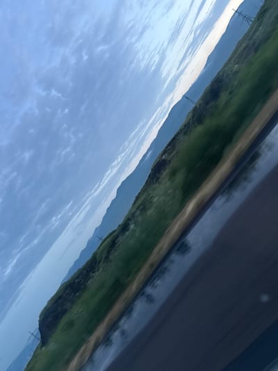 The image is a slightly angled, motion-blurred view of a rural landscape, possibly taken from a moving vehicle. The sky is a pale blue with scattered, wispy clouds. In the middle ground, a green, grassy hillside rises sharply, with several power line towers visible on its crest. In the background, layers of hazy blue mountains recede into the distance. The foreground is dominated by a dark, reflective surface, likely a wet road or body of water, showing reflections of the sky and landscape. There are no people or specific urban features to pinpoint a precise location within Hrazdan, Armenia, but the general characteristics suggest a mountainous, somewhat undeveloped area.