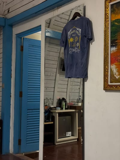 The image depicts an indoor scene, likely a guest room, located in Toyapakeh, Indonesia.
On the left, a light blue louvered door is set within a white frame, revealing a lighter interior beyond. The adjacent wall is constructed from horizontally aligned, light-colored wooden planks. Centrally positioned is a tall, vertical rectangular mirror. A dark blue t-shirt with a white graphic design is suspended on a metal hanger hooked over the top edge of this mirror. The t-shirt's graphic features the text "NUSA CENINGAN VIBES BALI," "LET'S SUNSET," symbols of suns, and two tigers.
The mirror's reflection shows a section of a room with a wall made of light-colored horizontal wooden planks. In front of this reflective wall, a light-toned wooden cabinet structure is visible. On its top surface are various objects, including several clear plastic bottles, a black electric kettle, a green bottle, and other miscellaneous items. A silver-colored mini-fridge is recessed within an open compartment of the cabinet below the top surface.
To the right of the mirror, a plain white wall is visible, on which an ornate gold-framed abstract painting is mounted, featuring prominent orange, green, and yellow hues. The floor throughout the visible area is a dark reddish-brown. No people are present, and no overt actions or interactions are occurring.