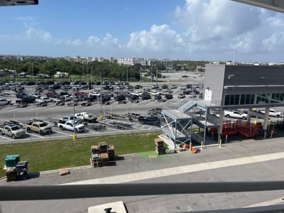 A high-angle, full shot captures a vast parking lot filled with trucks and trailers under a partially cloudy blue sky in an urban environment. The lot is organized, with vehicles parked in designated spots. In the foreground, a modern, grey, multi-story building with large windows dominates the right side of the frame, featuring an outdoor staircase. Beyond the parking lot, a line of trees separates it from a distant residential area with light-colored, multi-story buildings and palm trees. Several stacks of wooden pallets and a green dumpster are visible on the paved ground to the left. A metal railing is present in the bottom portion of the image. The overall scene depicts a transportation hub or a large parking facility.