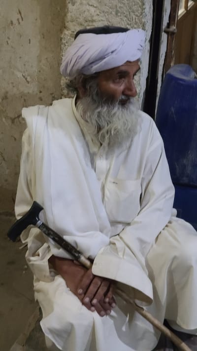 An elderly man lies on his side on a rough, pale surface. He wears a white turban and a long white robe. His long white beard is prominent. A dark walking cane rests between his folded hands. A blue container is visible in the background. The location is Ziarat, Pakistan.