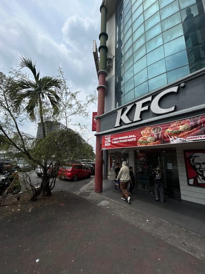 This outdoor, vertical-format image captures a street scene in front of a KFC restaurant within the Subang Jaya City Council, Malaysia. The setting includes a modern building with large, reflective glass panels on the left, reflecting parts of the cloudy sky.
The main subject is a KFC establishment, prominently featuring a large "KFC" sign on the building facade. A red banner hangs by the entrance, displaying images of fried chicken, burgers, fries, and drinks, alongside text that reads "DAPATKA" (likely part of "DAPATKAN" meaning "Get it") and "THE ONE BOX LEBIH BERSELERA, LEBIH PUAS HATI!" which translates to "THE ONE BOX MORE DELICIOUS, MORE SATISFYING!".
Several individuals are visible. One person in a light hoodie and brown pants walks towards the KFC entrance, while another in a dark outfit is seen further inside the restaurant. More people are partially visible inside.
A sidewalk made of paving stones runs along the building. Beyond the sidewalk, a street or parking area is visible with a red car and other vehicles. The scene is framed by a large palm tree on the left and other leafy trees on the right, indicating a green environment. The sky is overcast with light gray clouds, suggesting a cloudy day. The overall activity appears to be routine commercial foot traffic. No specific emotions are discernible from the individuals.