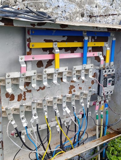 The image shows an outdoor electrical distribution box, likely in Marhi, India, due to the rustic and weathered appearance. The box is made of metal and shows signs of rust and peeling paint, particularly around the edges and on the interior back panel. Inside, a complex arrangement of electrical components is visible.

Three horizontal busbars, painted blue, yellow, and pink, run across the top of the box. These busbars are supported by silver metal brackets. Numerous electrical wires, varying in color (black, blue, yellow, white, and what appears to be a green-sleeved cable), are connected to various points within the box. Some wires are secured with metal clamps, while others are terminated with what seem to be screw-on connectors. Tape, likely electrical tape, is used to secure some of the wire connections.

A prominent object on the right side of the box is a circuit breaker. It is a grey rectangular unit with a black handle and a label that reads "Insul-Guard" and "CE". The circuit breaker has several terminals and wires connected to it.

The interior of the box appears somewhat disorganized, with wires running in various directions. In the lower part of the image, more wires are seen exiting the box and leading downwards, where they are surrounded by green vegetation. The overall environment suggests an outdoor setting, possibly near a wall made of rough, textured material, with some greenery growing around the base of the electrical box. There are no people visible in the image. The scene depicts standard, albeit aged, electrical infrastructure.