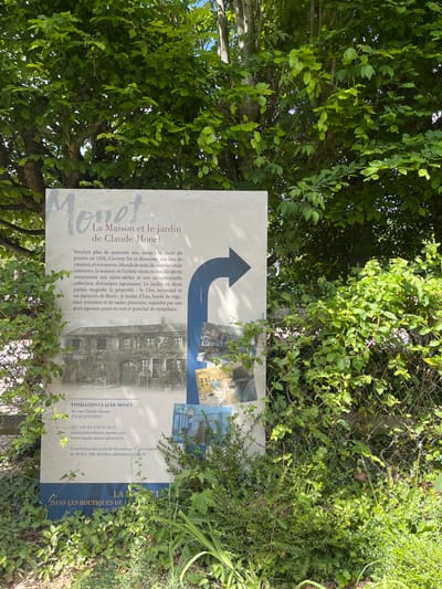 A sign stands partially obscured by foliage in a garden setting. The sign is a white rectangular panel with black lettering.  It advertises "La Maison et le jardin de Claude Monet" in Giverny, France (latitude: 49.075, longitude: 1.53).  The text describes Monet's home and garden, a popular tourist destination. The sign also contains contact information for "Fondation Claude Monet,"  which includes an address, phone number, email, and website.   The sign includes a large black arrow pointing to the right.  There are small black and white photographs of Monet's home and garden surrounding the sign. The text in French reads: “For over forty years, Giverny was his residence, his source of inspiration, the place where his creative work, "Monet's World of Sens," unfolded and took root.  His home, a place of family life and a refuge of memories, is now a museum.  It features an exceptional collection of Japanese prints. The garden, in two distinct parts, magnifies the property: The Clos normand and the oriental garden. This garden includes impressive Japanese willow trees, flowerbeds, a stream, and a Japanese bridge painted green and studded with water lilies.”  The background of the image is a lush green garden with foliage obscuring the sign. The sign is likely located in the garden of Monet's home in Giverny.