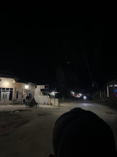 The image depicts a street scene at night in Landuh, Indonesia. The perspective suggests the photo was taken from a moving vehicle, likely a motorcycle, as a dark shape, possibly a helmet or shoulder, occupies the foreground on the right. The street is paved and illuminated by the headlights of a motorcycle in the distance and lights from buildings.
Several buildings are visible on the left side of the street, some appearing to be under construction or in a state of disrepair. One building shows a white facade with open window frames and visible interior structures. A motorcycle is parked in front of this building. Further down the street, more lights indicate other structures and possibly more activity.
On the right side of the street, the road surface is visible, reflecting the ambient light. The overall impression is a quiet, residential or semi-commercial area at night, with minimal pedestrian activity. The time of day is clearly nighttime due to the darkness and artificial lighting. There are no visible signs of text in the image. The atmosphere is subdued, typical of a nocturnal urban or semi-urban environment.