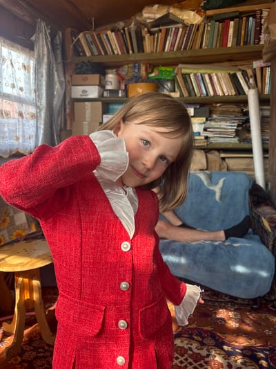 A young girl with blonde hair and blue eyes stands in a cluttered room, wearing a red jacket with white ruffles. She is positioned in the foreground, facing the camera with her head tilted and her right hand near her face. Behind her, bookshelves filled with books and various items line the walls. A blue blanket with a white bird-like pattern covers a sofa on which a person's legs are visible. The room appears to be a storage area or a casual living space. The lighting suggests it is daytime, with sunlight streaming in from a window to the left. The overall impression is of an indoor, informal setting. The specific location in the city of Маркидоновка, Russia is not determinable from the visual information in the image.