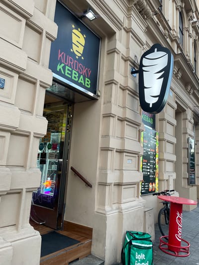 A street-level exterior shot captures a KORDSKY KEAB shop located in Brno, Czechia. The shop features a prominent black sign with a stylized yellow kebab graphic and the text "KURDSKY KEBAB" in pink and green lettering. To the right of the entrance, a black sign shaped like a rotating kebab is mounted on the building's facade. A menu board displaying food items is posted next to the kebab sign. In front of the shop, a red Coca-Cola branded table with a bicycle parked nearby is visible. A green Bolt Food delivery bag rests on the pavement. The building's architecture suggests an older European style with decorative stonework.
