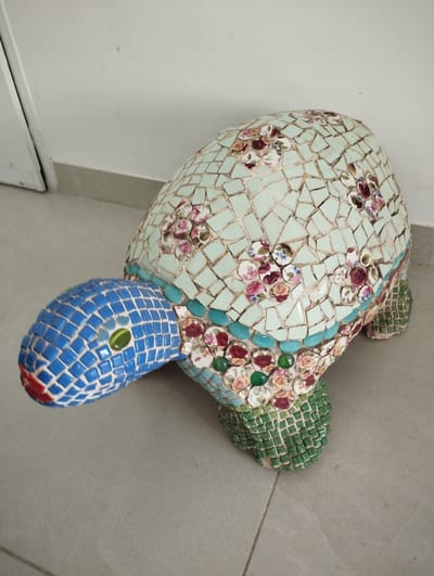 A mosaic statue of a turtle is positioned on a tiled floor against a white wall. The turtle's head and neck are covered in blue mosaic tiles with a red tip and one green eye. Its shell is predominantly light blue mosaic tiles with a pattern of small, colorful floral designs. The edges of the shell are adorned with a band of turquoise mosaic tiles and more floral mosaic pieces. The turtle's legs are covered in green mosaic tiles, giving them a textured appearance. The statue is a decorative object, likely placed indoors or in a covered outdoor area, given the tiled floor and wall. The lighting suggests an indoor setting, possibly during the daytime due to the bright illumination. There is no visible text in the image. The location context indicates this is in Pardes Hana-Karkur, Israel.