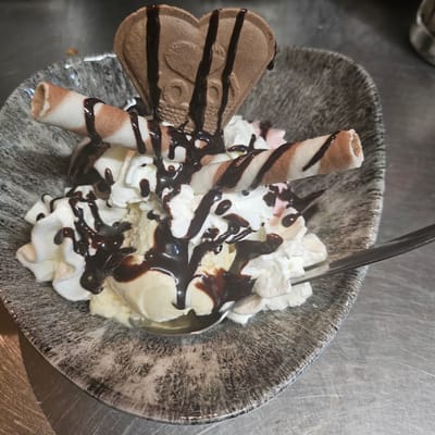 A high-angle shot depicts a dessert in a speckled gray bowl. The dessert consists of a scoop of light-colored ice cream, whipped cream, chocolate syrup, a striped wafer roll, and a chocolate-covered wafer cookie shaped like a butterfly. A spoon is inserted into the ice cream. The bowl rests on a stainless steel surface. The location is unspecified, but given the prompt, it could be assumed to be in Fehmarn, Germany.