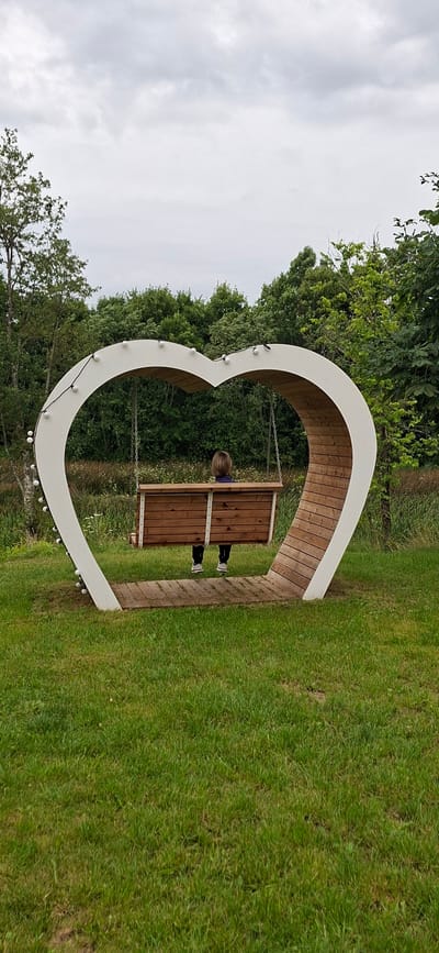 A person is sitting on a wooden swing shaped like a heart, facing away from the camera. The swing is suspended by chains from the top of the heart frame. The heart frame is white on the outside and lined with wood on the inside, with a string of lights decorating the outer edge. The swing and frame are situated on a grassy area with trees and vegetation in the background. The sky is overcast with clouds. The scene appears to be outdoors in a park or garden.