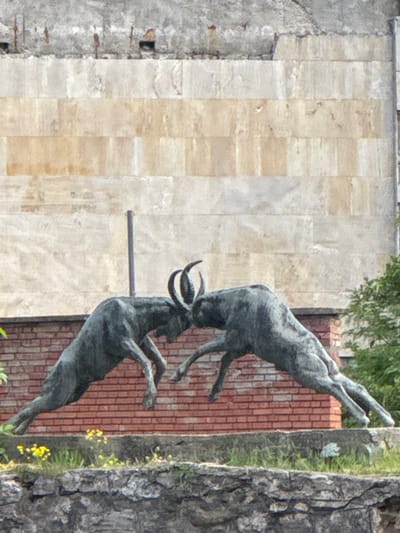 The image shows the sculpture "Twin goats" by sculptor Petar Lazarov in Rudozem, Bulgaria. Two bronze goats are depicted facing each other, with their horns almost touching. The sculpture is positioned in front of a brick wall and a stone wall covered with greenery. The background features a light-colored stone building.