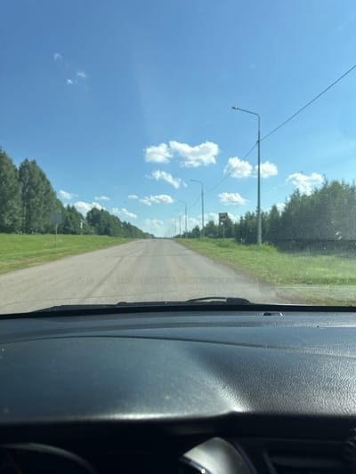 The image shows a view from inside a car looking out at a straight, paved road lined with trees on both sides. The sky is blue with scattered white clouds. There are streetlights visible along the side of the road. The dashboard of the car is visible in the foreground. The scene appears to be in a rural or suburban area. This image is consistent with the landscape of Verkhneyarkeyevo, Russia, a town located in the Republic of Bashkortostan, known for its natural beauty and agricultural surroundings.