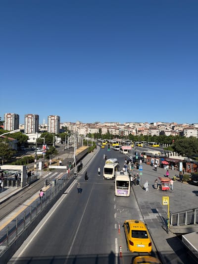 The image depicts a street scene in Bakırköy, Turkey, under a clear blue sky, suggesting it is daytime with good weather. The perspective is from an elevated viewpoint, looking down at a busy road and a city skyline.
Several vehicles are visible on the road, including yellow taxis, buses, and what appear to be smaller vans. People are scattered throughout the scene, walking on sidewalks, waiting at what looks like a bus or tram stop, and some are on the road. The architecture of the buildings suggests an urban environment, with a mix of older, multi-story buildings and some more modern, taller structures. Trees line parts of the street, adding some greenery to the urban landscape.
In the foreground, there is a tram track or similar rail system. In the midground, a yellow taxi with "TET 13" visible on its license plate is prominently featured. Other yellow taxis are also present. A sign with a yellow "P" in a blue circle indicates parking. Traffic lights are visible, with one showing a green light. There are also vending kiosks or small shops along the sidewalk.
The activity taking place is typical of a bustling urban environment: people moving about their day, vehicles traversing the road, and the general flow of urban life. The scene is well-lit, indicating it's likely mid-morning or afternoon.
Notable details include the clear blue sky, the presence of public transportation, and the density of urban development. The overall mood is one of everyday activity in a lively city. Some text is visible, including "TET 13" on the taxi, and a sign that appears to be a parking sign. There's also text on some of the buses, though it's not clearly legible.