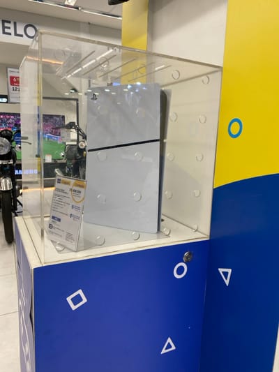 A white PlayStation 5 console is displayed vertically within a transparent acrylic security case. The console features a distinct black horizontal stripe and the 'PS' logo on its upper left side. The acrylic case sits on a pedestal with a blue base adorned with white outlines of PlayStation controller symbols (circle, square, triangle). A small lock mechanism is visible on the blue pedestal.
To the left of the console inside the case, a clear acrylic stand presents a white information card. The card prominently displays "Coppel" and "Contado $1.499.999", along with "CONSOLA SONY PS5 825GB BLANCO" and additional product specifications. This branding confirms the image's location as a Coppel retail store in Quilmes, Argentina.
The background shows a brightly lit retail environment. Reflections of overhead lighting are visible on the acrylic case. Other merchandise is partially visible, including a chrome headlight and tire of a motorcycle to the left, and a television screen displaying what appears to be a soccer match further in the background. Partial store signage with "ELO" and "Victory 100" can also be observed. No individuals are present in the foreground or clearly visible in the background.
