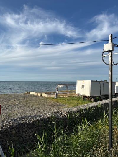 This image captures a bright, sunny coastal or waterfront scene, likely in Taman, Russia. The expansive blue sky, filled with long, wispy white clouds, dominates the upper portion of the frame. Below the sky, a wide body of calm water, possibly the Black Sea or Sea of Azov, stretches across the mid-ground to a distant horizon.

Along the water's edge, there's a constructed embankment featuring gabion baskets (wire mesh cages filled with rocks) forming a retaining wall, with loose gravel covering the adjacent ground. Behind this barrier, dense green reeds and tall grasses grow, particularly prominent on the right side of the image.

On the land, set back from the shoreline, a white, rectangular container-like structure is visible. It appears to be a portable building or utility unit, possibly mounted on a trailer, featuring a small window and a vent or exhaust on its roof. Part of another similar structure is seen behind it.

In the foreground, on the left, a dark utility pole or mast stands, fitted with a white rectangular box and black cables. A small, indistinct white object is also visible on or near the water's surface by the shore.

The scene is illuminated by clear daylight, suggesting late morning or early afternoon, and the weather is fair. There are no people visible, and no specific activities are taking place. The presence of gabions and portable structures indicates a utilitarian or possibly industrial aspect to this waterfront location. No legible text is discernible.
