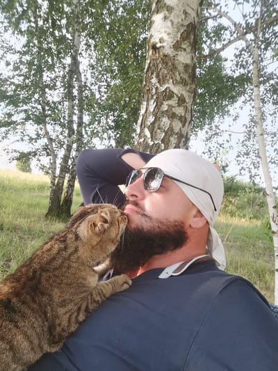 In this medium shot, a man with a dark beard and a white bandana on his head is leaning back against a tree. He is wearing sunglasses and a dark blue shirt. A tabby cat is nuzzling his face, with its paw resting on his chest. The man appears relaxed and affectionate towards the cat. The background consists of a grassy field with several birch trees, suggesting an outdoor setting. The lighting indicates it's daytime, likely late afternoon or early evening given the soft light and shadows. The location is Semypolky, Ukraine.
