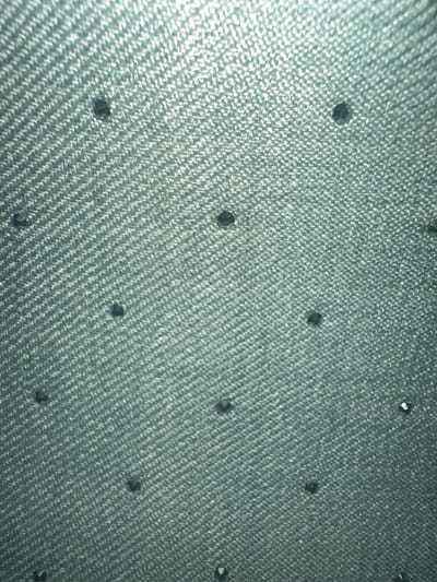 This media file is a close-up photograph of a textured, greenish-blue fabric, possibly denim, adorned with small, dark, faceted embellishments that appear to be rhinestones or beads. The fabric has a distinct woven pattern, with diagonal lines of raised texture. The embellishments are scattered unevenly across the surface, with some appearing more prominent and reflective than others, suggesting they catch the light. The lighting in the image is somewhat harsh, creating bright highlights on the fabric and the embellishments, and casting subtle shadows. The overall impression is of a decorative textile, likely a garment or accessory, viewed from a macro perspective. There is no discernible text, people, or specific environmental cues to suggest a particular time of day, weather, or outdoor location. The context of Letterkenny, Ireland, is provided externally, but the image itself does not contain visual information to confirm this setting.