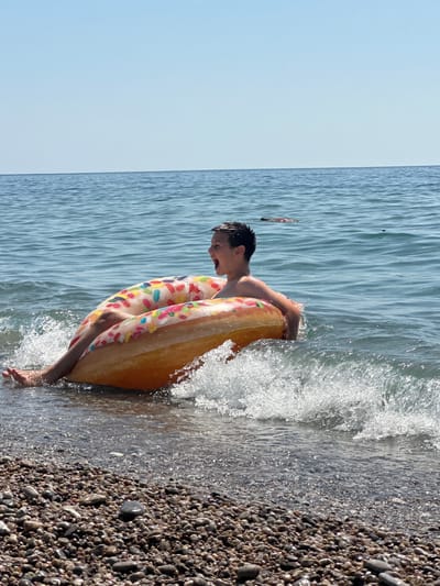 In the city of Đurovići, Montenegro, a young boy is in the water at the beach, sitting in a large, inflatable donut-shaped float. He is looking to his left with his mouth open, appearing to be shouting. The float is partially submerged in the water, with a small wave breaking around it, creating a splash. The water is calm and blue, with the horizon visible in the background. The beach in the foreground is covered in pebbles.