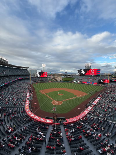 Here is a description of the image:

The image presents an elevated view of Angel Stadium, located in Anaheim, California, United States. The stadium is filled with spectators, most of whom are seated. The baseball field is visible with base lines, pitcher's mound, and home plate clearly delineated. Scoreboards display team logos and advertisements. A backdrop of mountains is faintly visible in the distance. Above the stadium, the sky is filled with clouds.