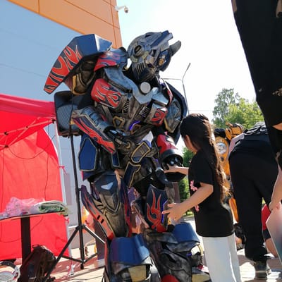 The image captures an outdoor event in Трубачеевка, Russia, during bright daylight hours under a clear, light blue sky. The central focus is a person in a large, highly detailed costume resembling Optimus Prime from the Transformers franchise. The costume features intricate blue, grey, and red armor plating with red flame-like decals and emits blue light from its eyes and chest/waist areas, simulating a metallic appearance.
To the right of the robot, a young girl with long dark hair, wearing a black t-shirt with a pink flower graphic and white pants, extends her right hand to touch the robot's forearm. Partially visible to the far right are sections of another individual's black clothing and legs, and behind the girl, another person in black attire leans over a yellow object that appears to be the upper section and head of a Bumblebee robot costume, identifiable by a visible round headlight.
On the left side of the frame, a red pop-up canopy is erected. Underneath the canopy, a small black table holds a clear plastic water bottle and some folded white and green fabric. A dark brown backpack rests on the ground near the table. A black speaker on a stand is positioned between the red canopy and the robot costume. In the background, a modern building with horizontal orange paneling features a white security camera mounted on its exterior. A streetlamp and green leafy trees are also visible further in the background. The ground is a paved surface.