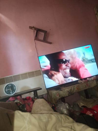A television displays a close-up of a Black man with sunglasses, smiling widely, with another person's arm around him, against a blurred background. The television is on a stand covered with a floral tablecloth. To the left of the TV is a white radiator. A round analog clock with a black frame and white face, showing the time as approximately 11:55, is mounted on the wall above the radiator. A thin black cable hangs down from the wall, connected to an unseen device. The room appears to be a bedroom, with a bed covered in various blankets and pillows in the foreground. The walls are painted pink and there is a light source creating a shadow on the upper left wall. There are no specific indicators of the location being Fanchykovo, Ukraine.
